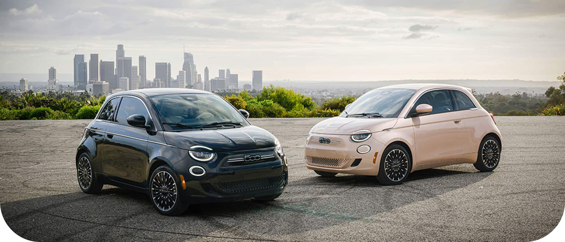Two Fiat 500e models—on the left ,a black 2024 Fiat 500e Inspired By Music and on the right, a rose gold 2024 Fiat 500e Inspired By Beauty—parked near a waterfront, with a sprawling skyline of high-rise buildings in the background.