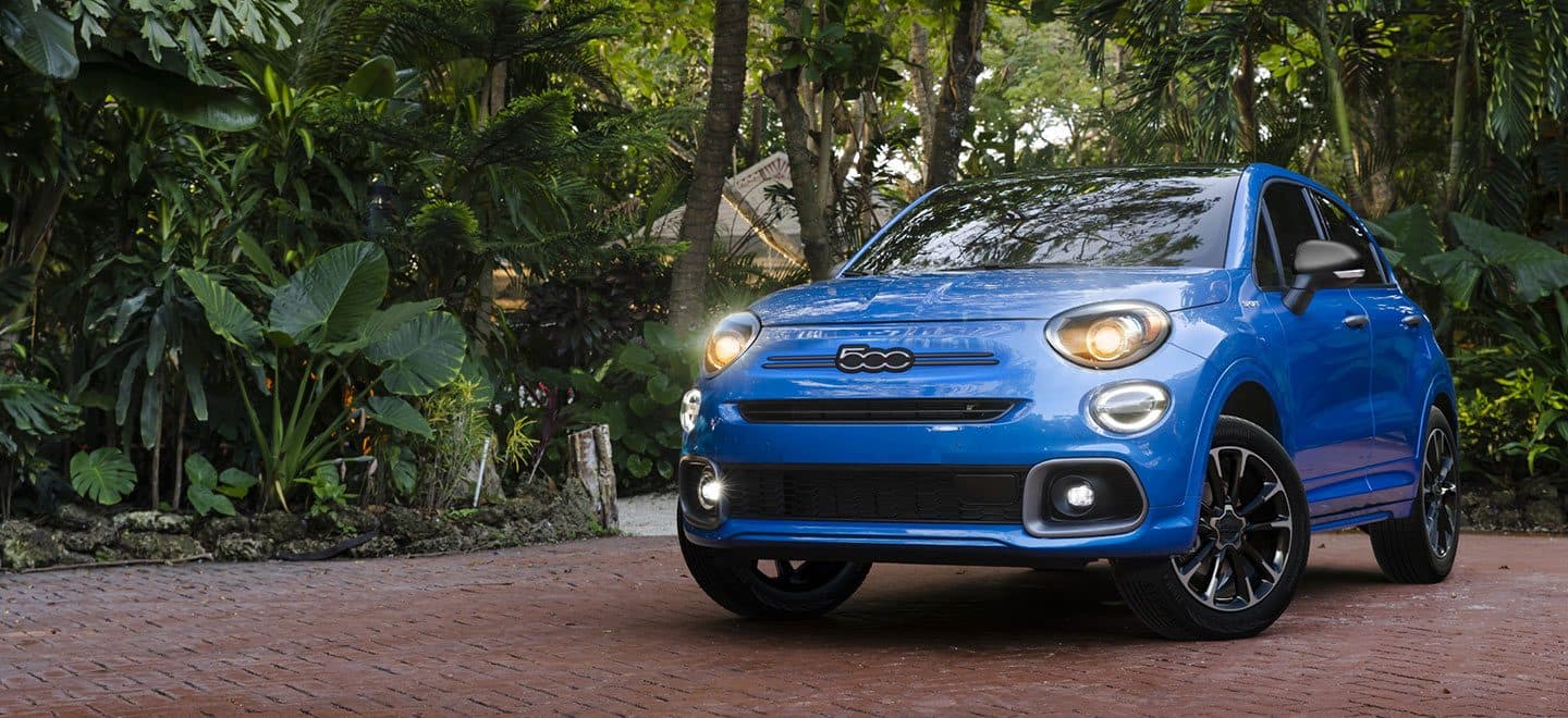 Display A blue 2022 Fiat 500X Sport with its headlamps on, parked in a residential neighborhood beside a lush garden.