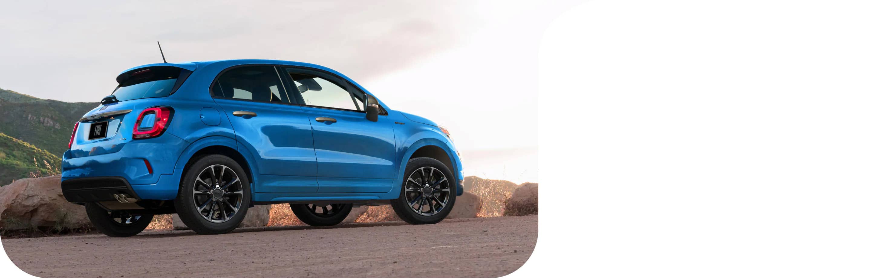 Vista trasera de medio perfil de un Fiat 500X Sport 2022 azul estacionado al costado de una carretera al lado de una montaña.