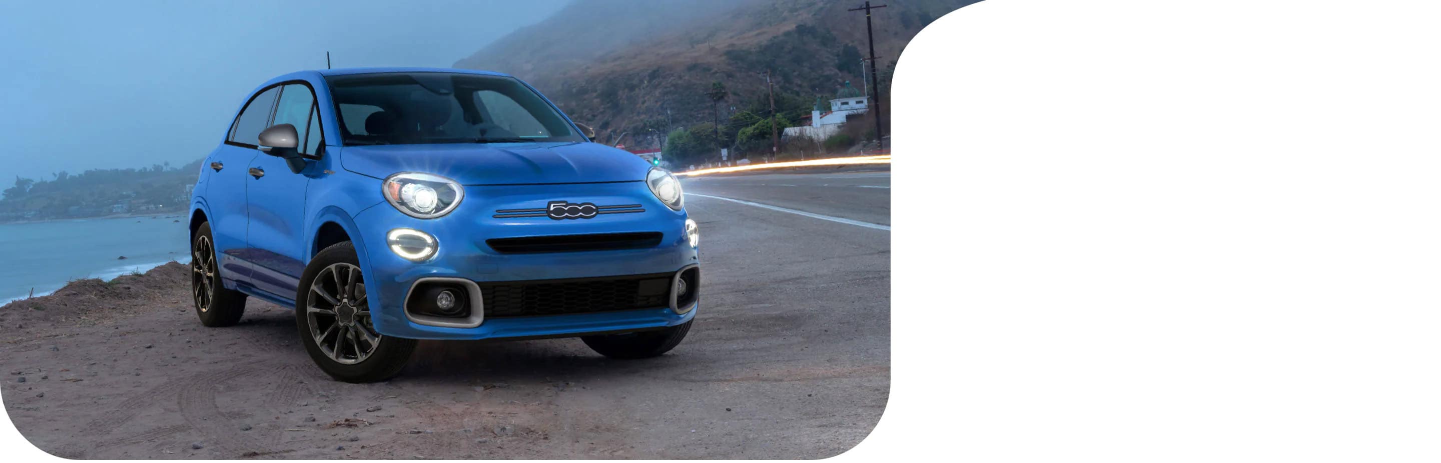 A blue 2022 Fiat 500X Sport with its headlamps on, parked on the shoulder of a highway beside a mountain lake at dusk.