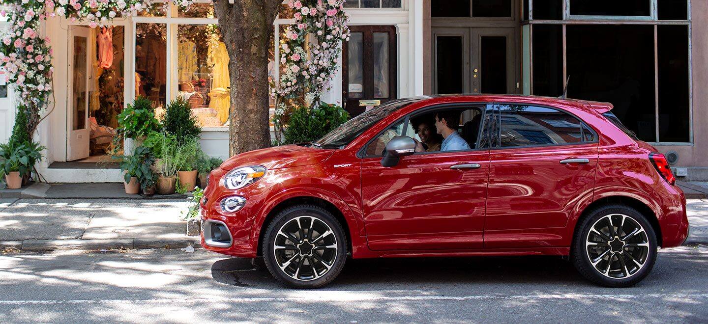 Display A profile of a red 2023 Fiat 500X Sport parked on a city street with shops in the background. A couple is seated in the front seats.