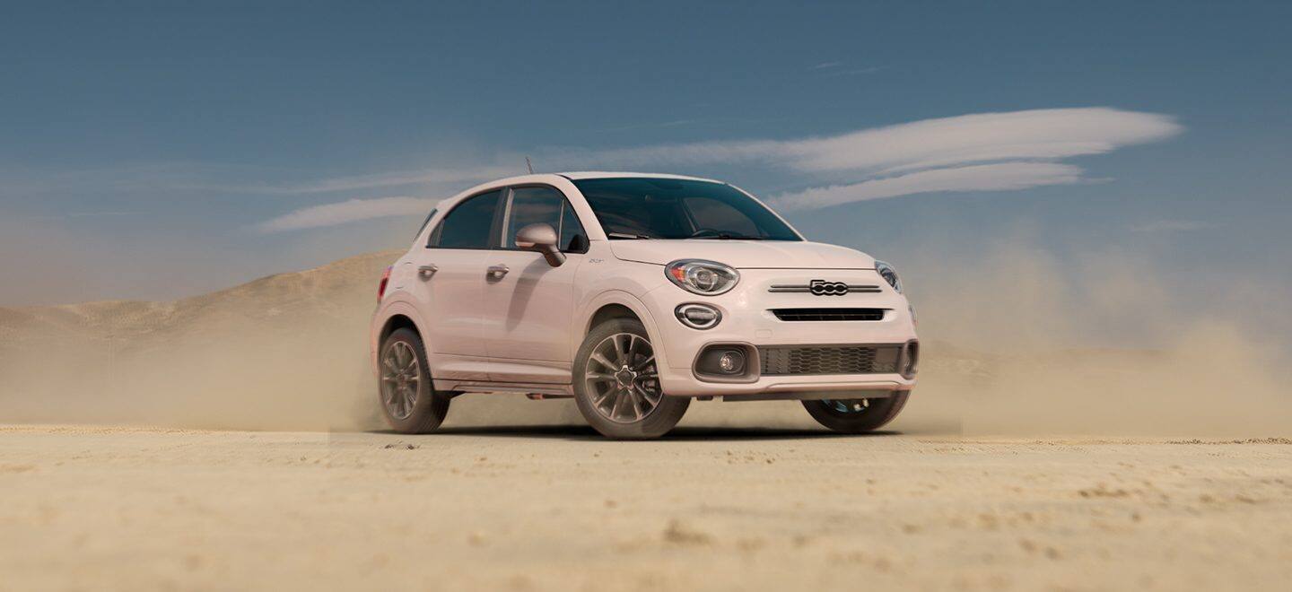 Display An angled profile of a white 2023 Fiat 500X Sport parked on sand with hills in the background.