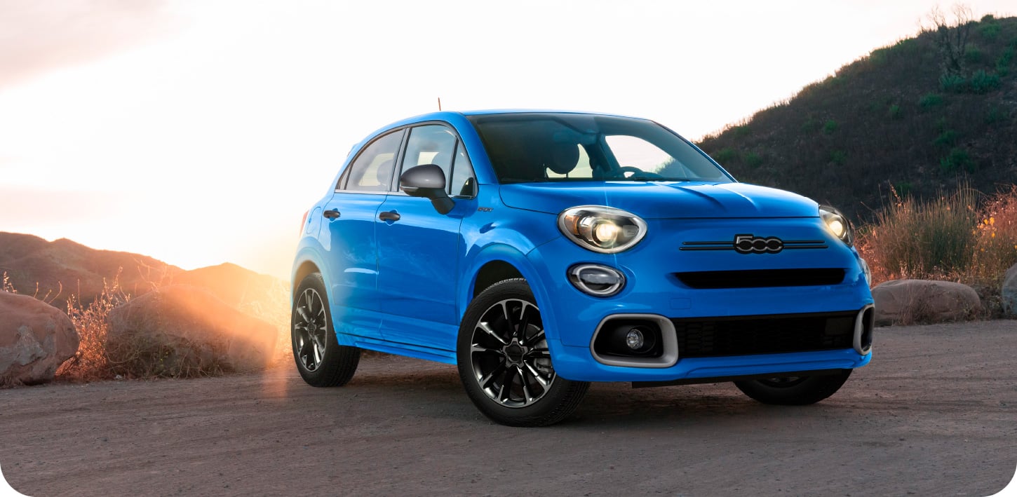 A blue 2023 Fiat 500X Sport parked on a clearing with the sun rising over hills in the background.