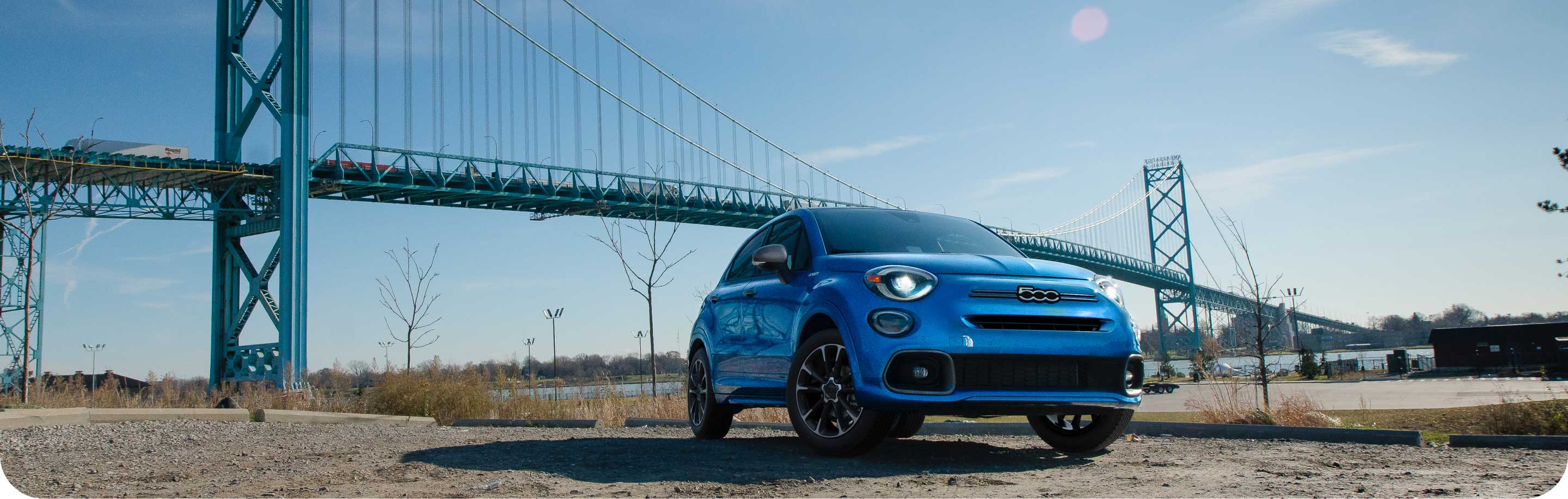 A blue 2023 Fiat 500X Sport parked on a clearing with the sun rising over hills in the background.