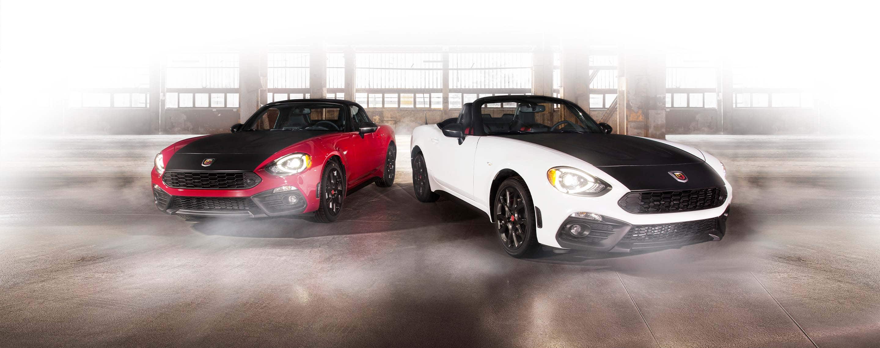 A red 2020 Fiat 124 Spider Abarth and a white 2020 Fiat 124 Spider Abarth with a black hood and its top down, side-by-side in a parking garage.