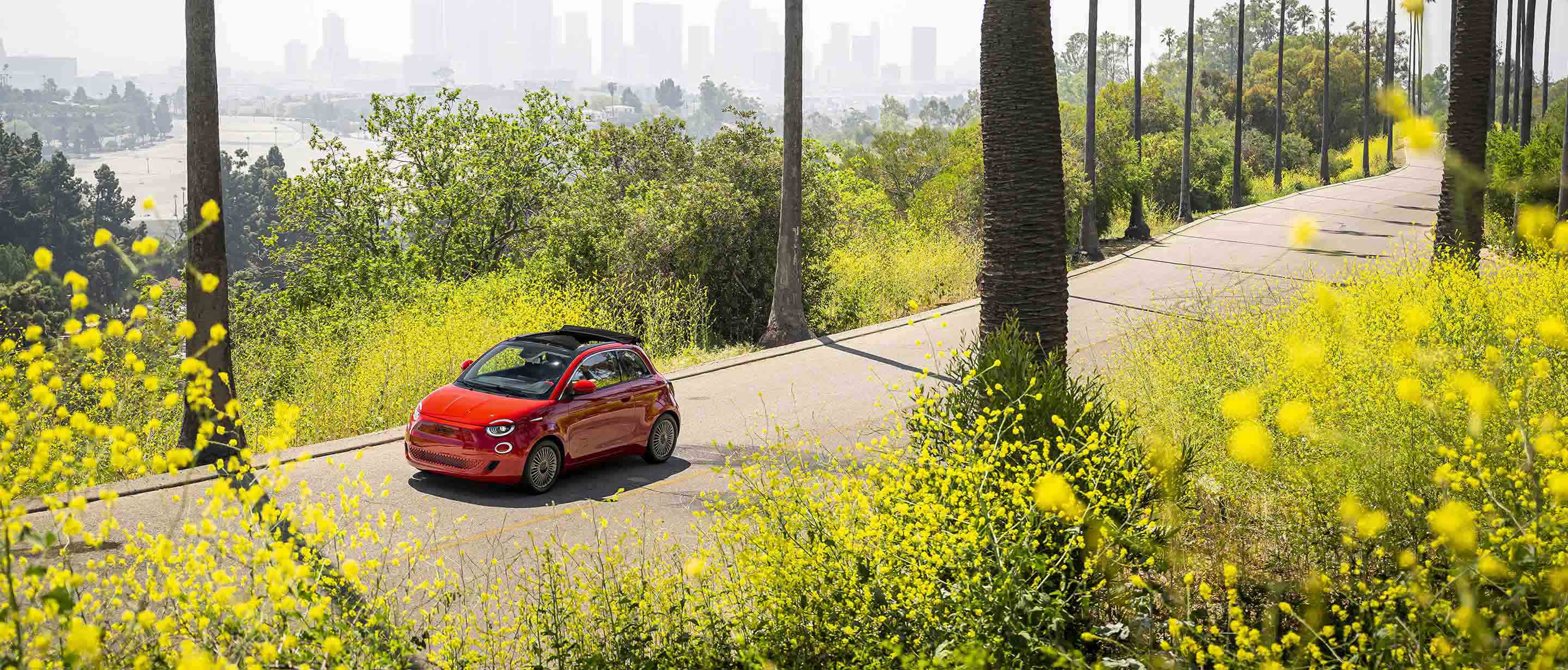 A red European model Fiat 500e being driven on a road surrounded by trees and bushes, with a river and high-rise buildings in the distance.