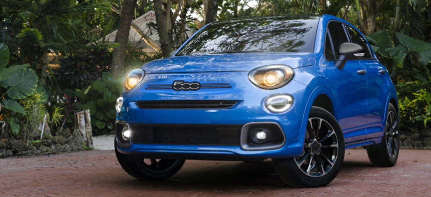 Display A blue 2022 Fiat 500X Sport with its headlamps on, parked on a residential neighborhood beside a lush garden.