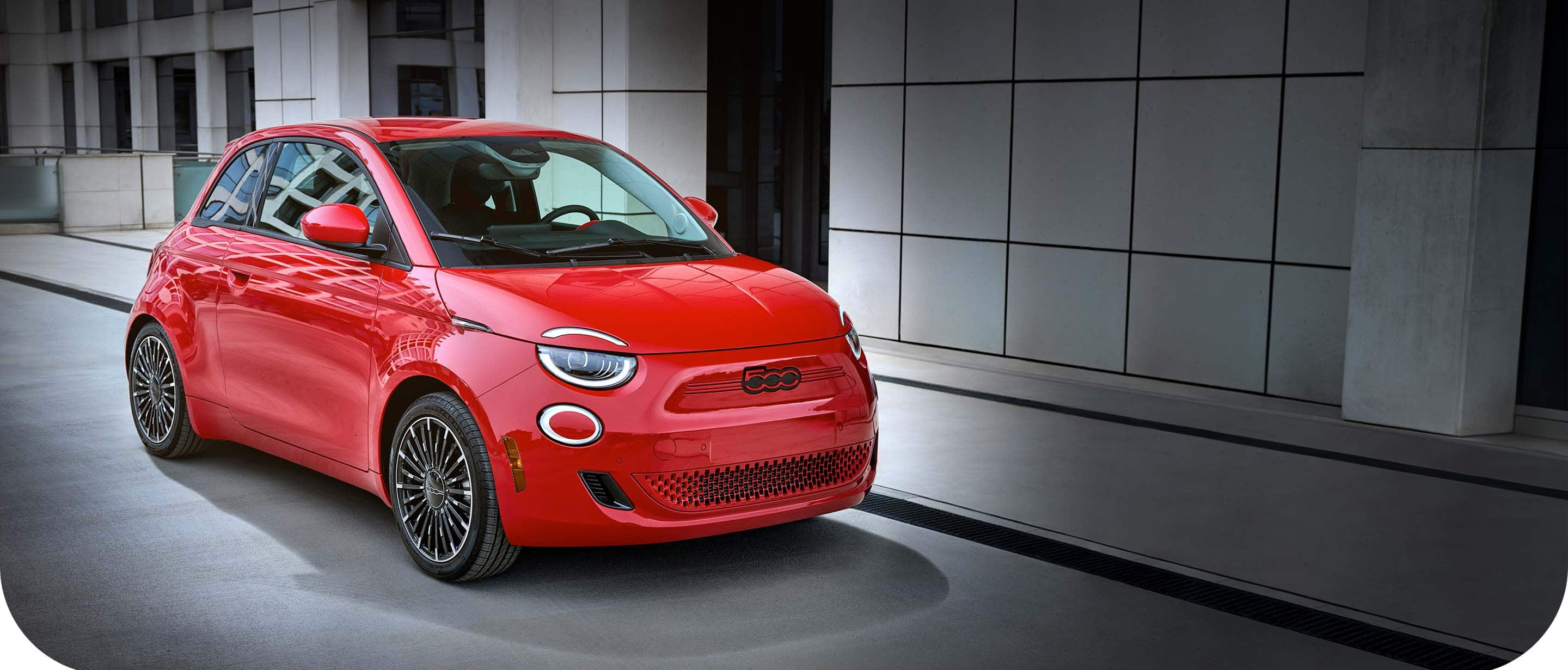 An angled front passenger-side profile of a red 2024 Fiat (500e) Red Edition parked beside a contemporary building.