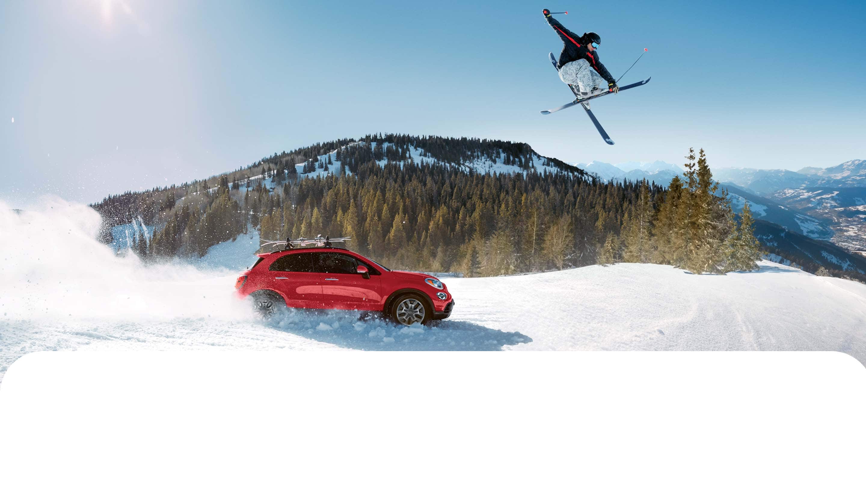 A passenger-side profile of a red 2022 Fiat 500X Trekking with aftermarket equipment, being driven through heavy snow, kicking up the white powder as it goes, while a skier jumps overhead and mountains rise in the background.