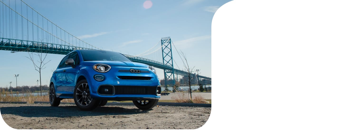 A blue 2022 Fiat 500X Yacht Club Capri parked in a clearing with a suspension bridge in the background.