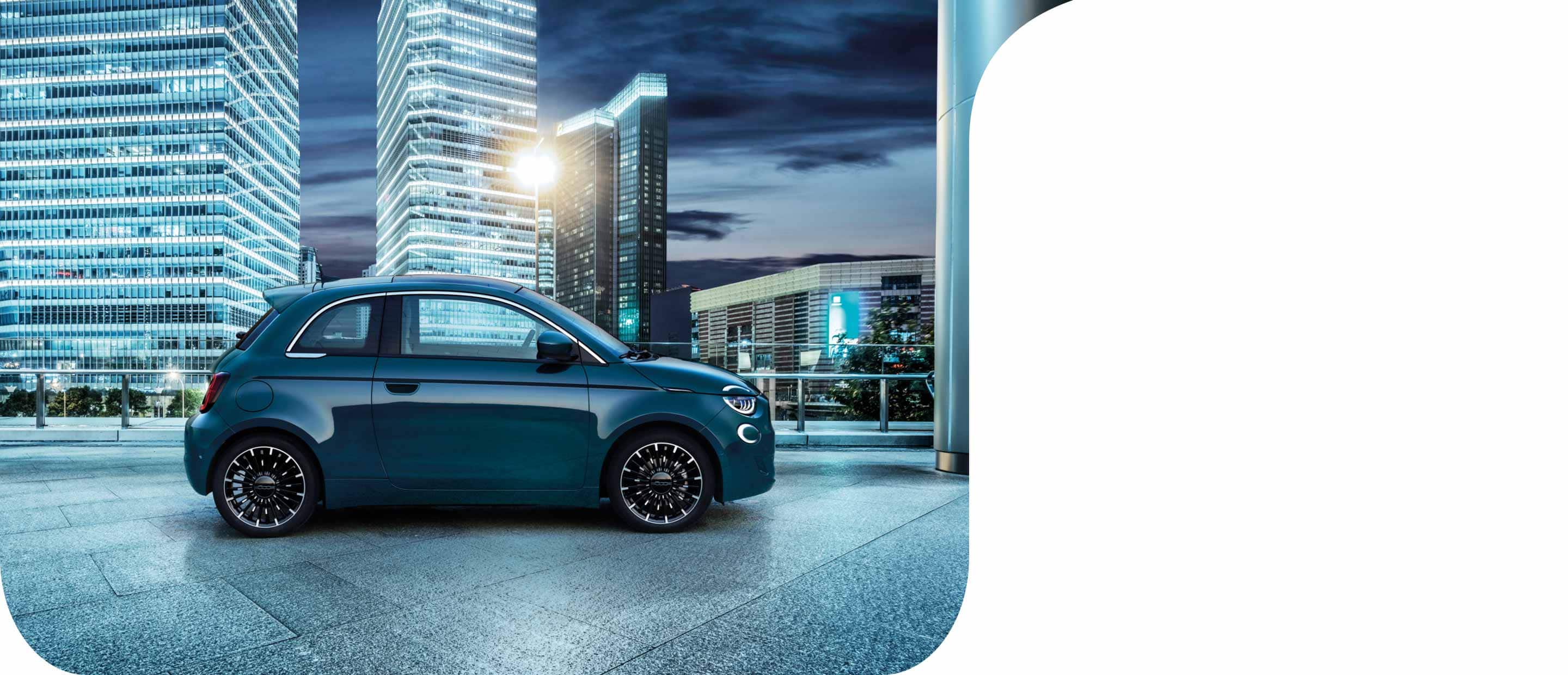 Perfil del lado del pasajero del modelo europeo del Fiat 500e Cinquecento 2023 azul estacionado en una estructura de estacionamiento en la azotea por la noche, con varios rascacielos de fondo.