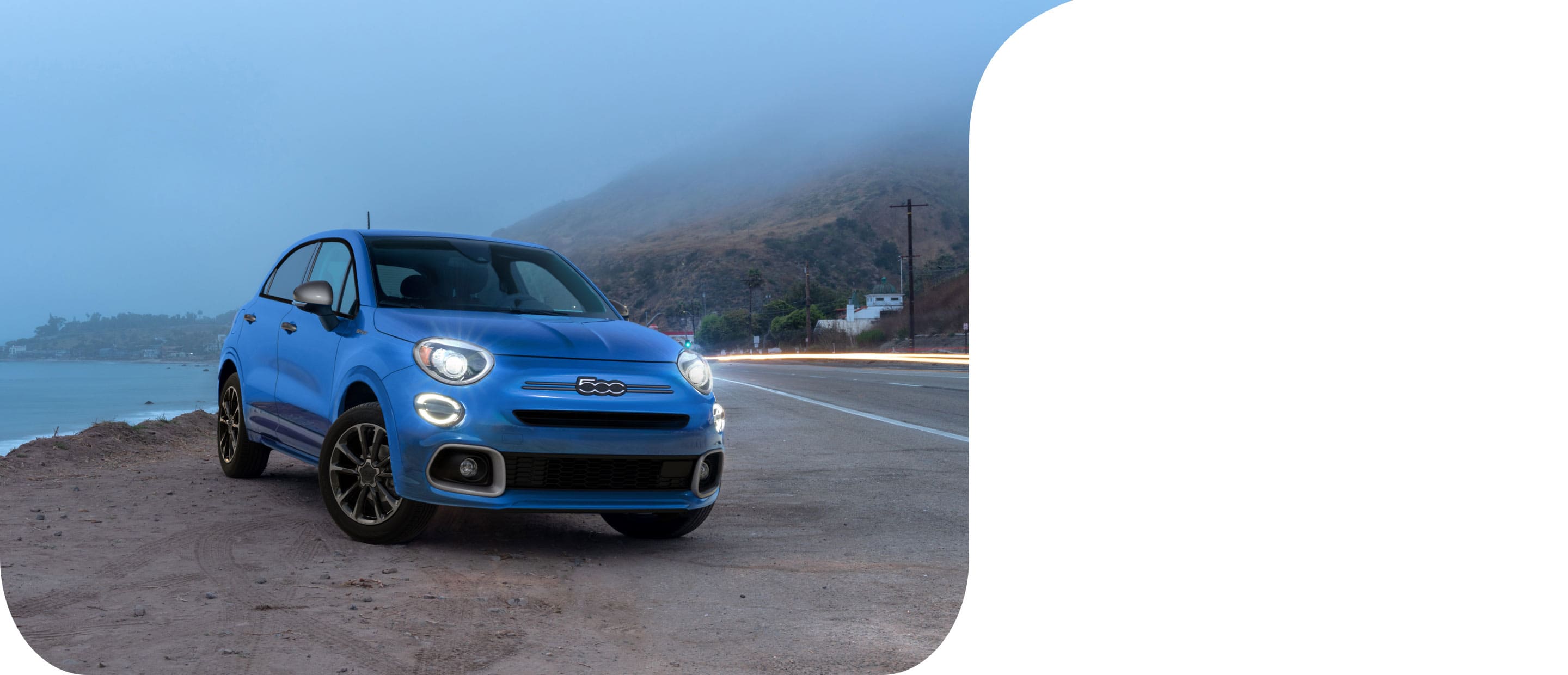 An angled front of a blue 2022 Fiat 500X Sport parked at the waters edge with mountains wrapped in fog in the background.