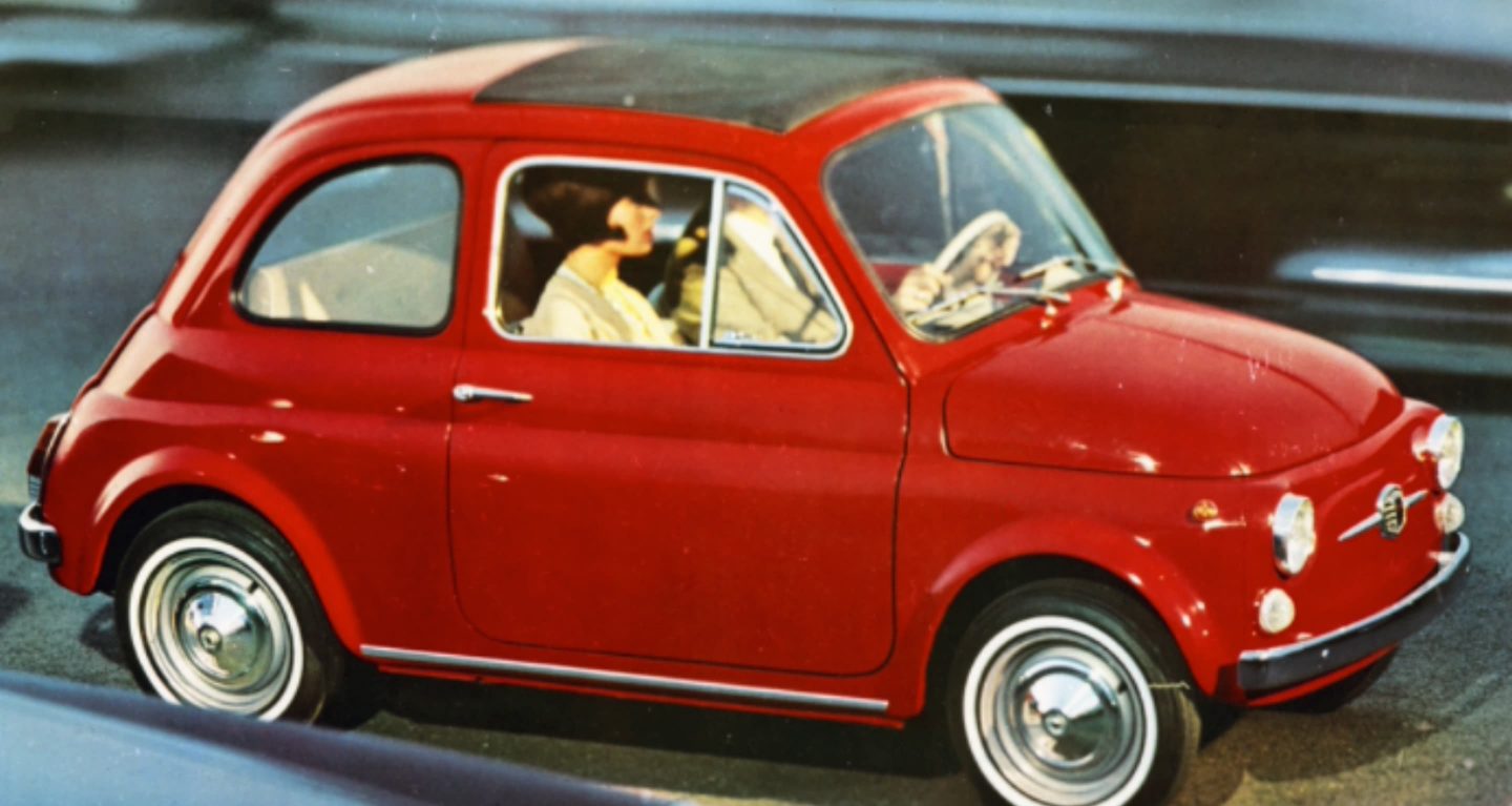 Display Wallpaper #4: A vintage photo of a classic Fiat 500 being driven on a street with a couple inside.