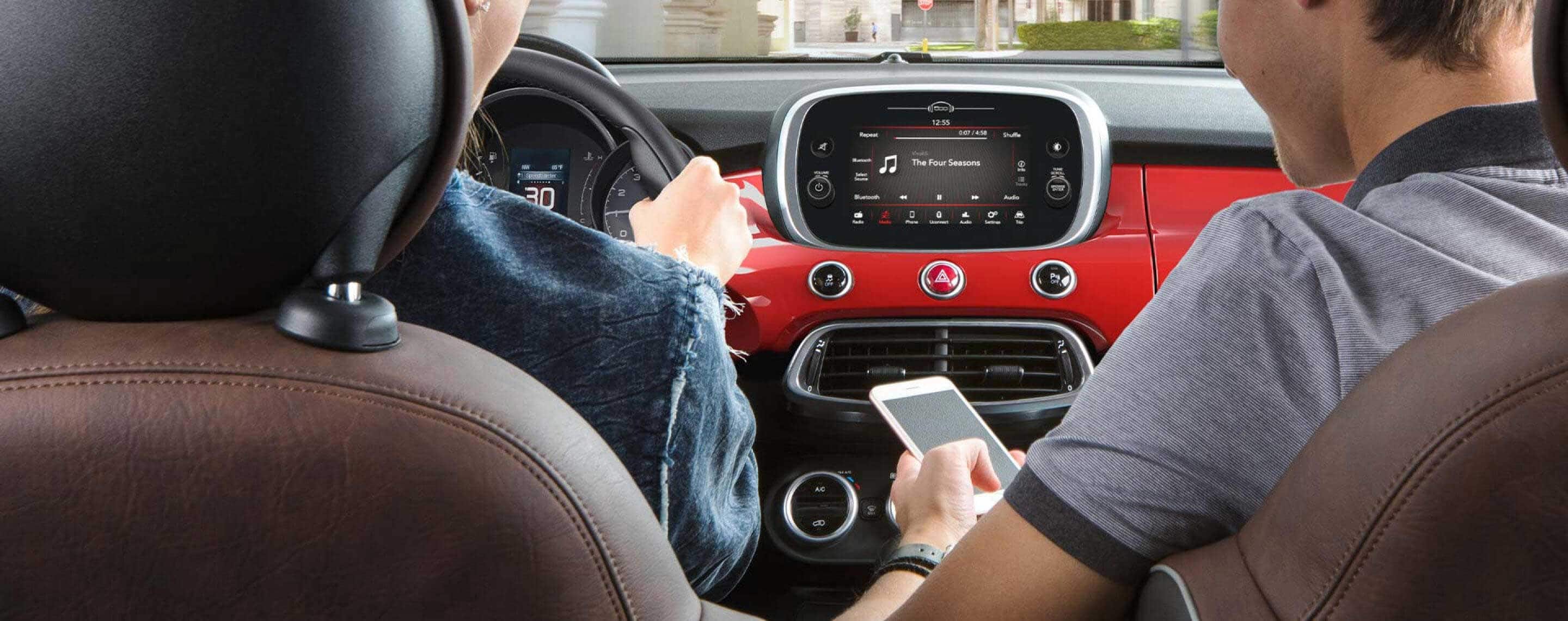 A man in the front passenger seat of a 2021 Fiat 500X Trekking Plus accessing features between his smartphone and the Uconnect touchscreen as the driver drives down a city street.