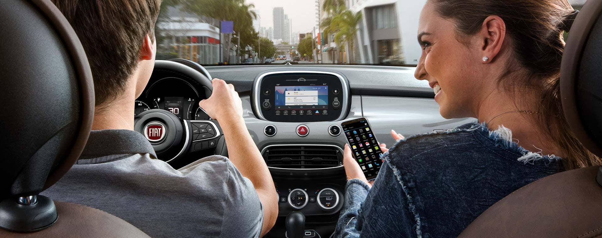 A woman in the front passenger seat of a 2021 Fiat 500X Trekking Plus accessing features between her smartphone and the Uconnect touchscreen as a man drives down a city street.