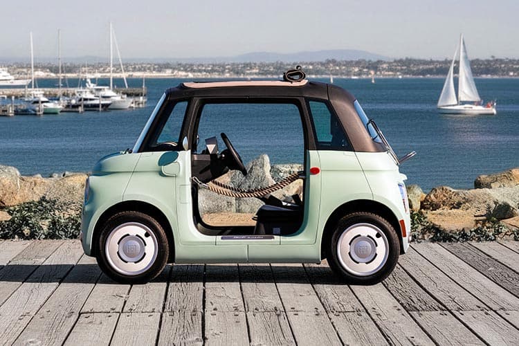 A mint green preproduction model Fiat Topolino Dolce Vita with door ropes and a rolled-up soft top, parked at a marina, shot in profile so you can see through the vehicle and beyond.