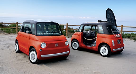 A Corallo Orange 2027 Fiat Topolino and a Corallo Orange 2027 Fiat Topolino Dolce Vita parked in a beachside lot.