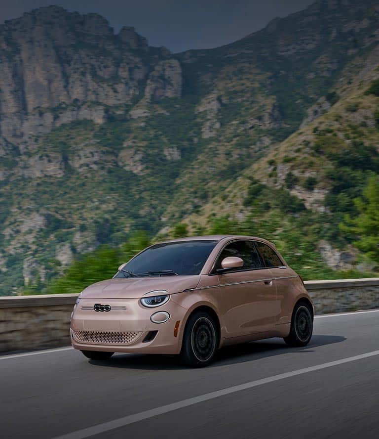 A rose gold 2025 Fiat 500e Icona traveling down a highway with mountains in the background.