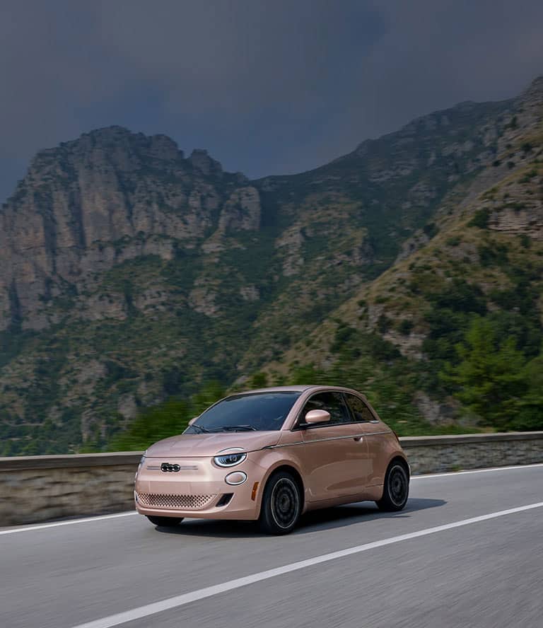 A rose gold 2025 Fiat 500e Icona traveling down a highway with mountains in the background.