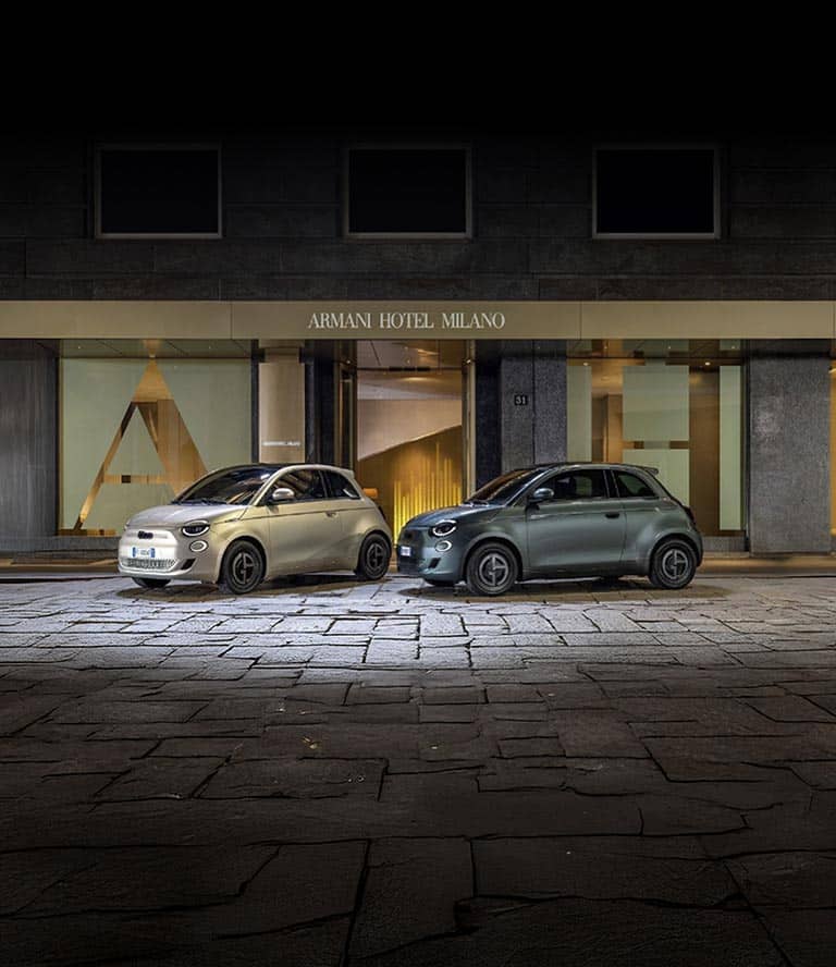 Dos modelos Fiat 500e 2025 Giorgio Armani Collector's Edition estacionados en una calle adoquinada. A la izquierda, un modelo en color beige grisáceo y a la derecha, un modelo en verde oscuro.