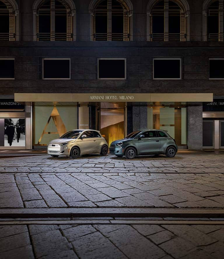 Dos modelos Fiat 500e 2025 Giorgio Armani Collector's Edition estacionados en una calle adoquinada. A la izquierda, un modelo en color beige grisáceo y a la derecha, un modelo en verde oscuro.