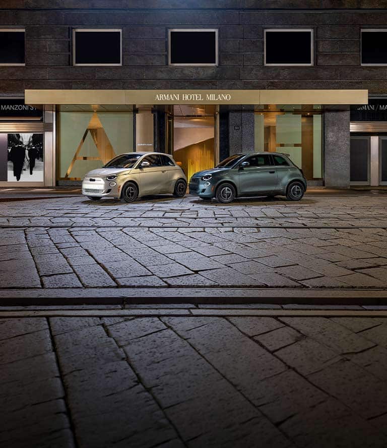 Two 2025 Fiat 500e Giorgio Armani Collector's Edition models parked on a cobblestone street. On the left a model in gray beige and on the right, a model in dark green.