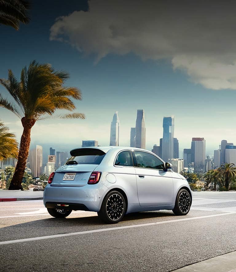 A passenger-side rear angle of an ice blue 2025 Fiat 500e Icona traveling down a highway with palm trees and a cityscape in the background.