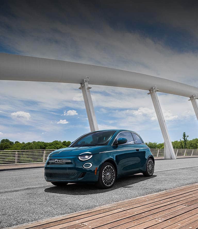 A driver-side front angle of a deep blue-green 2025 Fiat 500e Icona traveling on a bridge above the trees in the background.