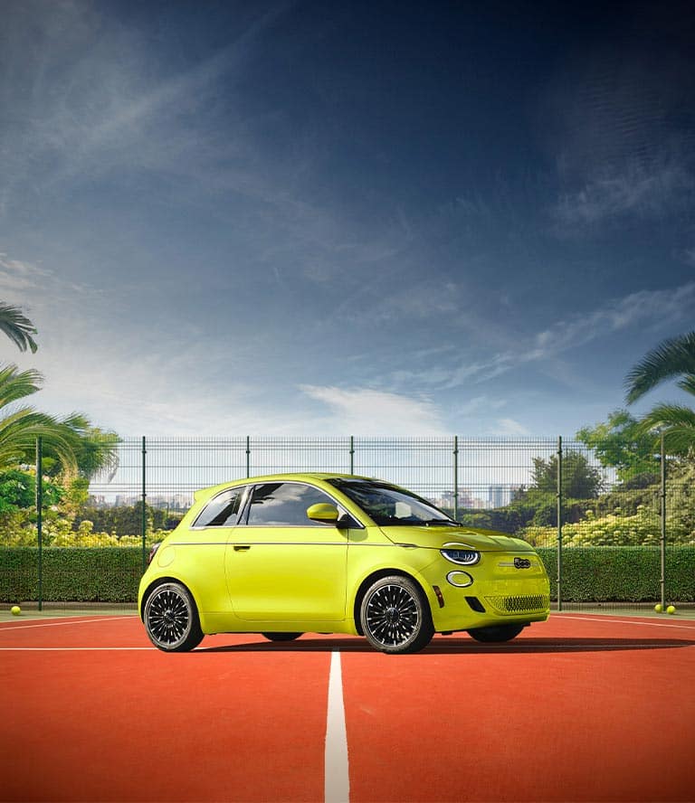 A passenger-side front angle of a 2025 Fiat 500e Icona in optic yellow (bright lime green with yellow undertones), parked on a tennis court.