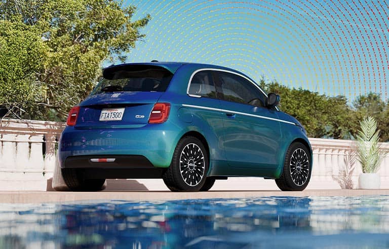 A rear passenger-side angle of a blue 2025 Fiat 500e Icona. The sky is filled with hundreds of tiny colorful zigzagged exclamation points.