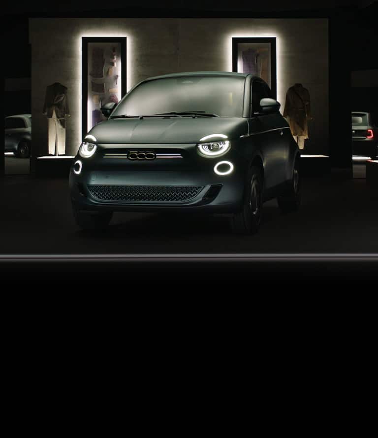 A green 2025 Fiat 500e Giorgio Armani Collector's Edition with a darkened high fashion display in the background.