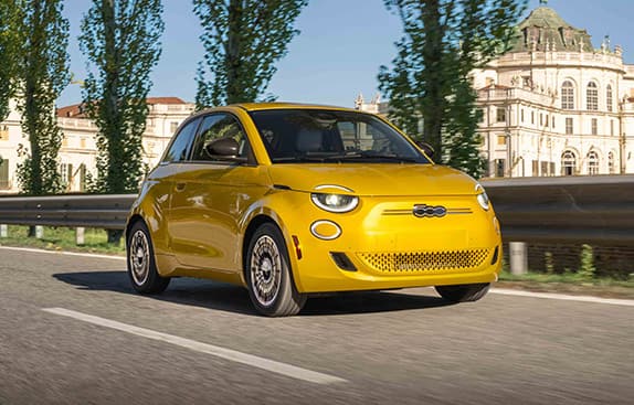 A yellow 2026 Fiat 500e Pop parked on a highway, with historical buildings in the background.