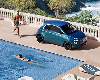 An overhead angle of a blue 2026 Fiat 500e Icona parked beside a pool, with a women swimming and a man walking toward the vehicle.