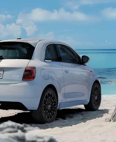 A white 2026 Fiat 500e Icona parked on a white sand beach at the water's edge.