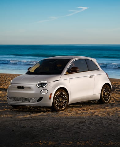 A white 2026 Fiat 500e Icona parked on a beach at the shoreline.