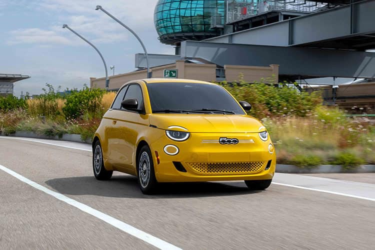 A yellow 2026 Fiat 500e Pop traveling down a highway with a building complex in the background.