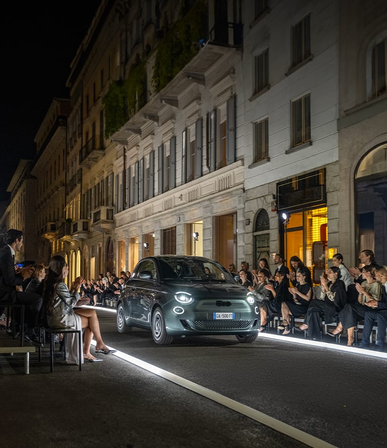 Ángulo delantero de un Fiat 500e Giorgio Armani Collector's Edition 2025, circulando por una calle preparada como pasarela iluminada y pasando a un grupo de admiradores.