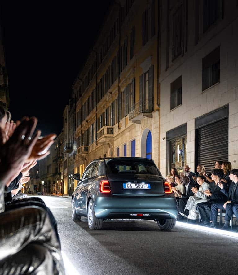 Ángulo trasero de un Fiat 500e Giorgio Armani Collector's Edition 2025 circulando por una calle preparada como pasarela iluminada y pasando a un grupo de admiradores.
