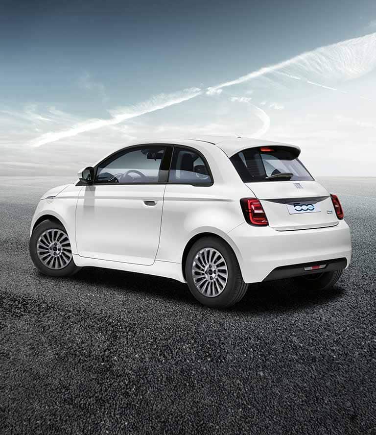 An angled driver-side rear profile of a white European model 2024 Fiat 500e parked on a gravel surface.