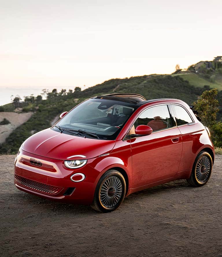 An angled driver-side front profile of a red 2024 European model Fiat 500e Red with its sunroof open, parked on a clearing with mountains and the sea in the background.