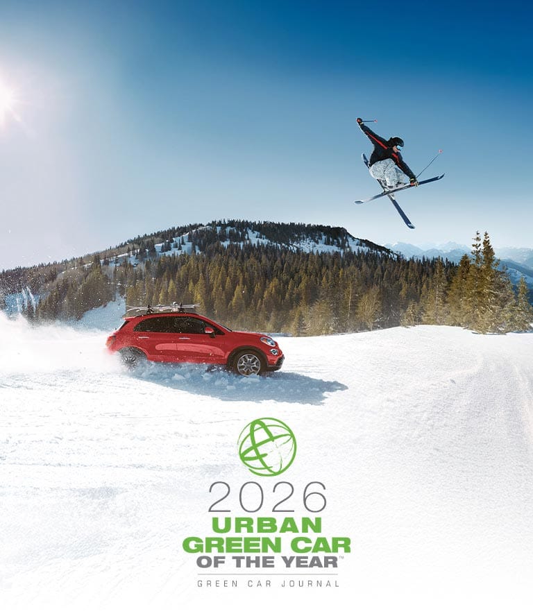 Perfil del lado del pasajero de un Fiat 500X Trekking 2022 rojo con equipamiento no original, circulando por un camino cubierto con mucha nieve, levantando polvo blanco mientras avanza, con un esquiador saltando por encima del vehículo e imponentes montañas de fondo.