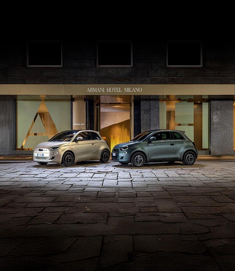 Two 2025 Fiat 500e Giorgio Armani Collector's Edition models parked on a cobblestone street. On the left a model in gray beige and on the right, a model in dark green.