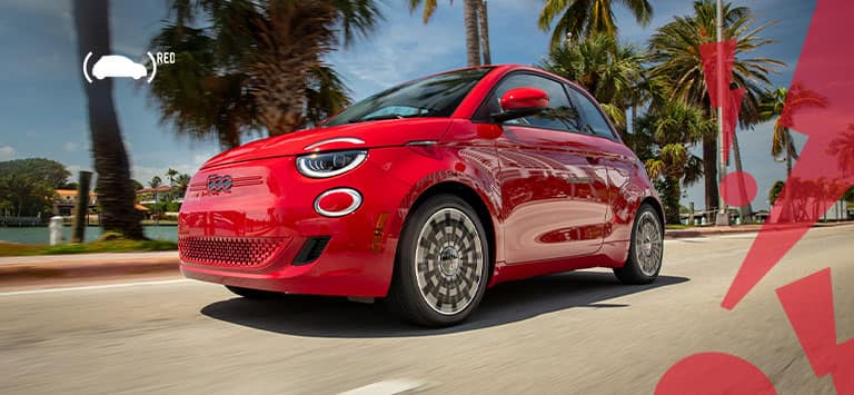 A front driver-side angle of a red 2025 Fiat 500e Red traveling on a highway beside palm trees and a body of water, with illustrated red zigzagged exclamation points in the right side foreground.