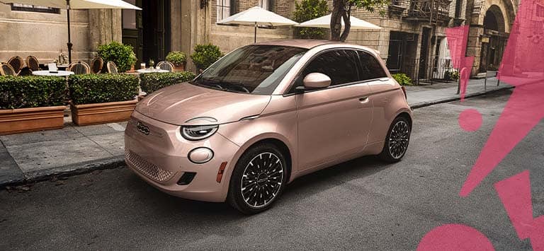 A front driver-side angle of a rose gold 2025 Fiat 500e Inspired by Beauty, parked on a city street beside a cafe, with illustrated hot pink zigzagged exclamation points in the right side foreground.