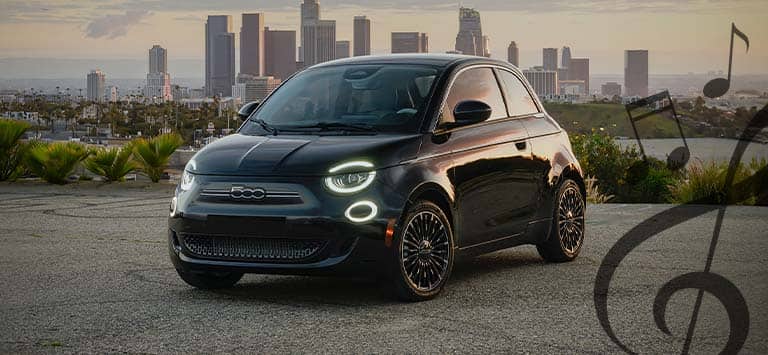 A front driver-side angle of a black 2025 Fiat 500e Inspired by Music with its headlamps on, parked on a clearing across the water from a bustling city at dusk and illustrated black musical notes in the right side foreground.