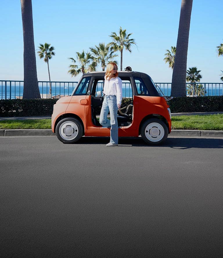 A woman stands at the doorway of an orange 2027 Fiat Topolino Dolce Vita that's parked on a street by a beach with palm trees.