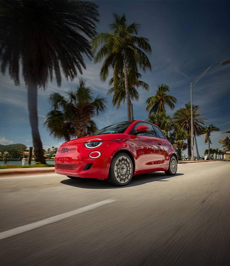 A red 2025 Fiat 500e Inspired traveling down a waterfront highway.
