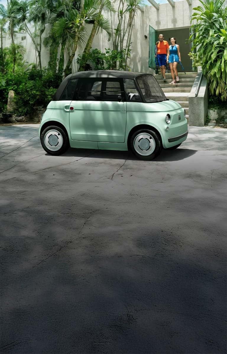A passenger-side profile of a light green Fiat Topolino parked in front of a tennis club, with a couple exiting the club.