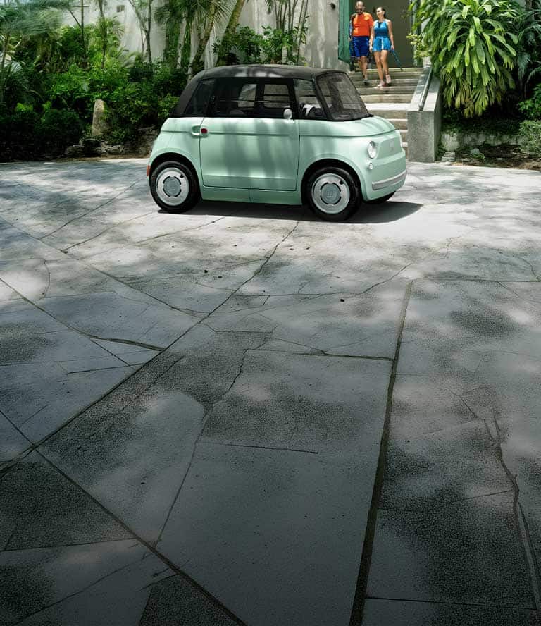 Vista de perfil del lado del pasajero de un Fiat Topolino verde claro, estacionado en frente de un club de tenis, con una pareja saliendo de dicho club.