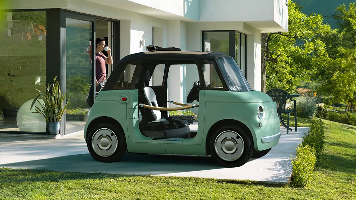 A mint green European model Fiat Topolino Dolce Vita with door ropes, parked in the driveway of a contemporary home.
